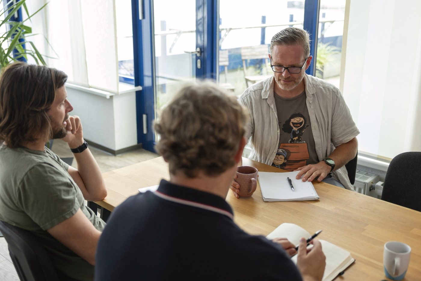 Drei Kollegen im Gespräch am Tisch, mit Notizblöcken und Kaffeetassen